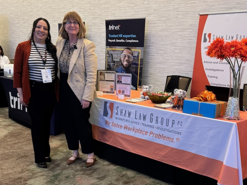 Two women pose at a Shaw Law Group booth with banners and a table of brochures and gifts in a conference setting.