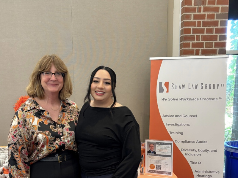Two women pose at a Shaw Law Group booth with banners and a table of brochures and gifts in a conference setting.