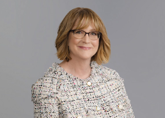Smiling woman with short blonde-brown hair and glasses, in a textured tweed blazer, for a professional headshot.