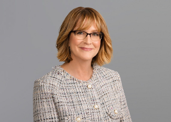 Head-and-shoulders portrait of a smiling woman with glasses, wearing a light tweed jacket against a gray backdrop.